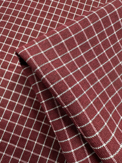 A close-up image of a maroon fabric with a white grid pattern. The high-quality Super Cheap Fabrics Linen Rayon Grid - Cabernet - 145cm is folded, showing multiple layers and the texture of the weave.