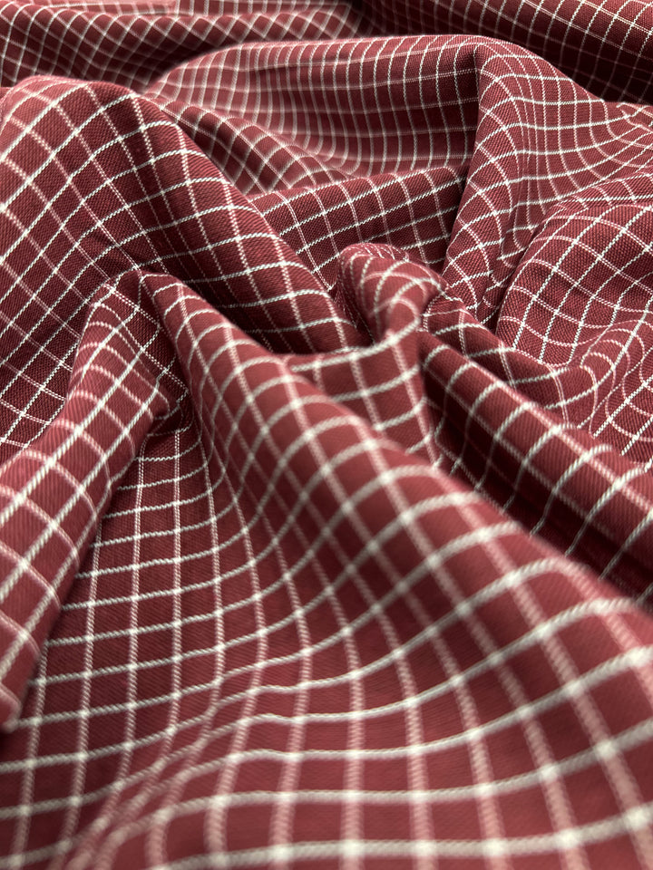 A close-up of **Linen Rayon Grid - Cabernet - 145cm** by **Super Cheap Fabrics** with a white grid pattern, showcasing intricate designs. The fabric is wrinkled and draped, creating folds and varying textures across the image.
