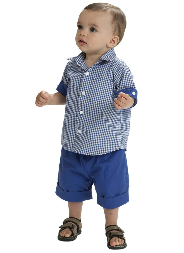 Dressed for a shopping trip, a toddler sports an outfit made from Super Cheap Fabrics' Pattern - Mccall's - M6016, featuring a blue and white checkered shirt with rolled-up sleeves and matching blue shorts. With short hair and arms slightly raised, he glances upward while wearing dark brown sandals. The simple white background enhances the charm of his poised anticipation.