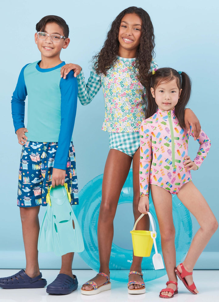 Three children, dressed in vibrant beach attire created using the Pattern - Mccall's - M8395 by Super Cheap Fabrics, stand beaming against a blue backdrop. One holds flippers, another clutches an inflatable ring, and the last proudly carries a sand bucket. They exude anticipation for an exciting day at the beach or pool.