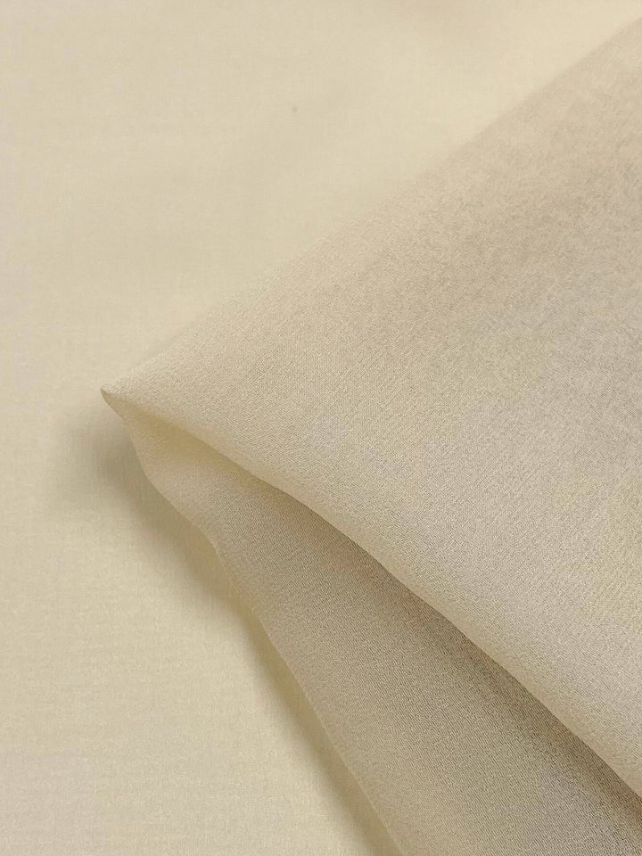 A close-up image of a piece of light beige fabric folded at one corner. The texture appears smooth and slightly sheer, suggesting that it might be Silk Georgette - Chardonnay from Super Cheap Fabrics. The background is blurred, focusing attention on the fabric, perfect for spring and summer outfits.