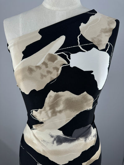 A close-up of a mannequin wearing a stylish, one-shoulder dress made from Super Cheap Fabrics' Printed Lycra - Brush Strokes - 150cm. The dress features a bold, abstract print in shades of black, white, beige, and gray, creating a sophisticated and modern look.