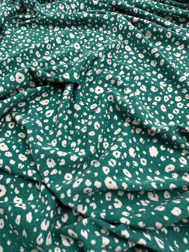 A piece of medium-weight green fabric with a white abstract dotted pattern is spread out, showing its texture and flowing folds. The pattern appears random and varies in size, adding a dynamic visual effect perfect for children's wear. This is the Printed Lycra - Dotty Green - 150cm by Super Cheap Fabrics.