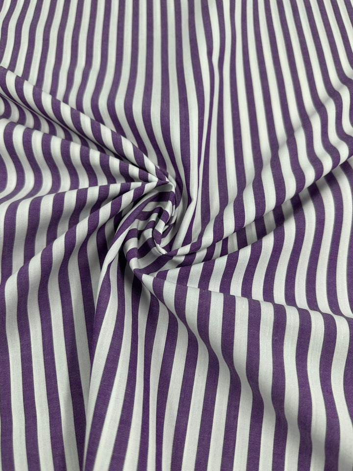 A close-up image of a purple and white striped fabric. The 100% cotton fabric, Cotton Lawn - Purple Stripe - 150cm by Super Cheap Fabrics, has fine vertical stripes and is slightly gathered in the center, creating a swirling pattern.