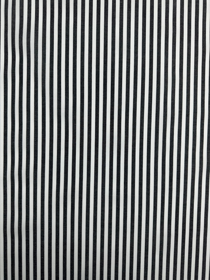 A close-up of a fabric featuring a pattern of alternating black and white vertical stripes. The evenly spaced stripes create a high-contrast design on this lightweight, 100% cotton material with a silky finish. This is the Cotton Lawn - Black Stripe - 150cm by Super Cheap Fabrics.