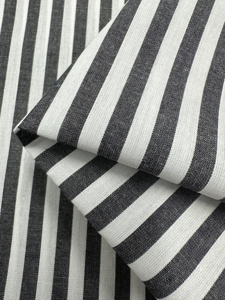 Close-up of black and white striped fabric with a folded section on top of a flat section. The lightweight fabric displays alternating vertical stripes, creating a classic and bold pattern. The 100% cotton texture of Super Cheap Fabrics' Cotton Lawn - Black Stripe - 150cm appears smooth with a slightly silky finish.