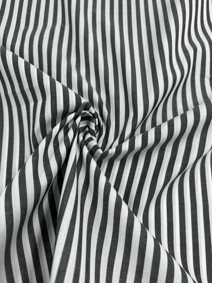 Close-up of a fabric featuring a white and dark gray vertical stripe pattern. The 100% cotton fabric is slightly gathered and twisted at the center, creating a rippled effect around the twist. The texture appears smooth with a silky finish and remains lightweight. This is the Cotton Lawn - Black Stripe - 150cm by Super Cheap Fabrics.