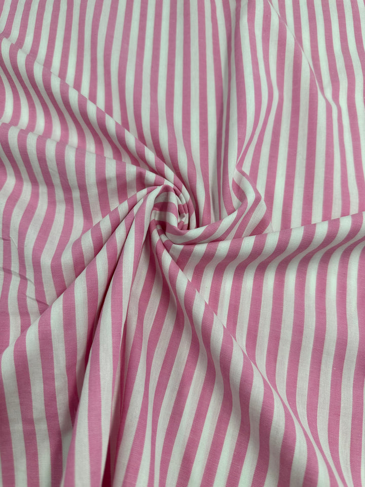 A close-up of a fabric featuring pink and white vertical stripes. The material appears to be Cotton Lawn - Pink Stripe - 150cm from Super Cheap Fabrics and is gathered in the center, creating a swirl pattern. The stripes are evenly spaced, adding to the fabric's orderly and symmetrical appearance.