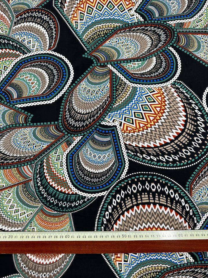 A close-up of *Super Cheap Fabrics' Textured Lycra - Africa - 150cm* featuring an intricate, colorful geometric pattern with overlapping paisley shapes. The design includes various zigzag and dot patterns in shades of blue, green, red, brown, and white. A ruler at the bottom measures the fabric in centimeters.