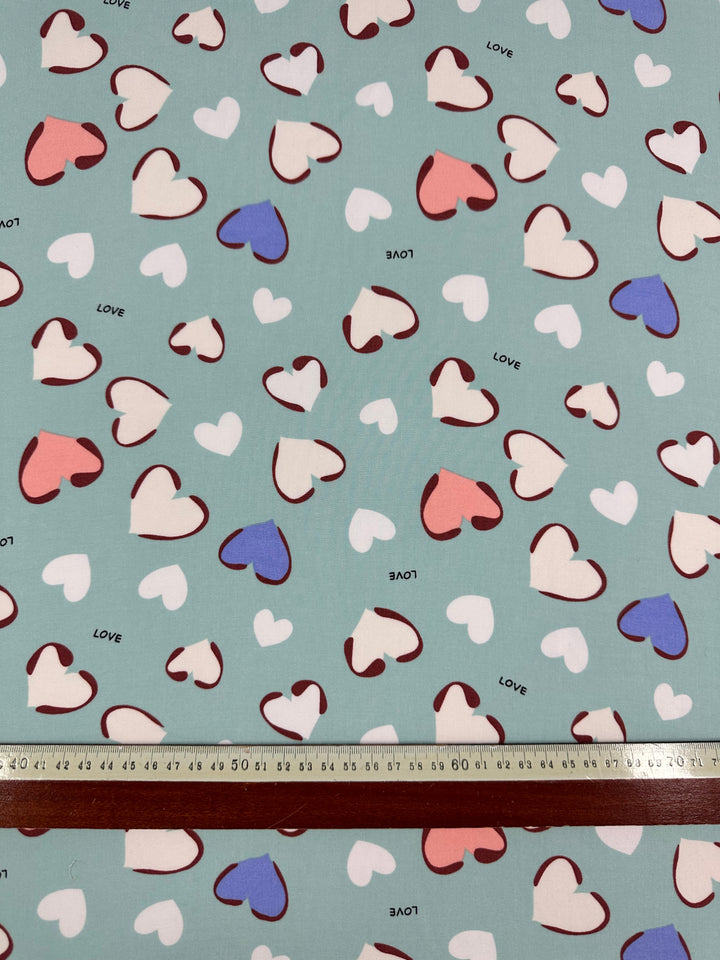 Introducing the Super Cheap Fabrics Printed Rayon - Love - 140cm: A fabric with a light blue background featuring a pattern of multi-colored hearts in white, pink, blue, and red outline. The word "LOVE" is scattered among the hearts. Perfect for hot weather, its lightweight drapey fabric will keep you cool. A ruler at the bottom shows the scale of the pattern.