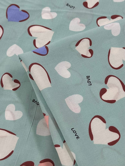 A close-up of a folded, light weight fabric with a light teal background. The fabric is decorated with white hearts, some outlined in red, and the word "LOVE" printed in black multiple times. There's a single blue heart among the white hearts, perfect for drapey clothing in hot weather. This is Printed Rayon - Love - 140cm by Super Cheap Fabrics.
