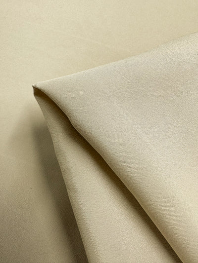 Close-up of beige, 100% polyester Microfibre - Oat - 150cm fabric from Super Cheap Fabrics with a smooth texture, shown with one corner folded over. The lightweight fabric's surface displays a slight sheen.