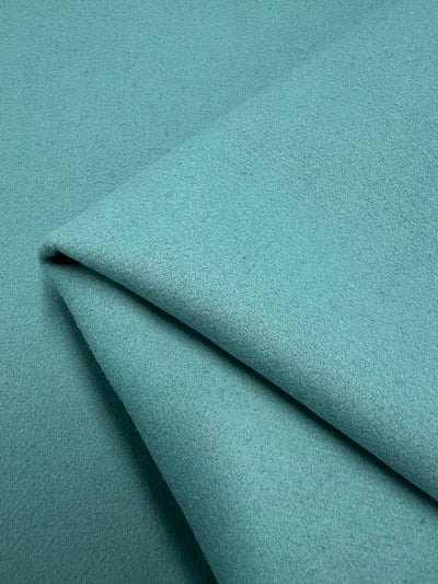 A close-up of the Wool Cashmere - Aqua Sea fabric by Super Cheap Fabrics, neatly folded. The texture is smooth and slightly fuzzy, indicating a soft material perfect for outer coats. The folds create subtle shadows and a gentle gradient in color, from light to slightly darker shades of aqua sea. Dry clean only.