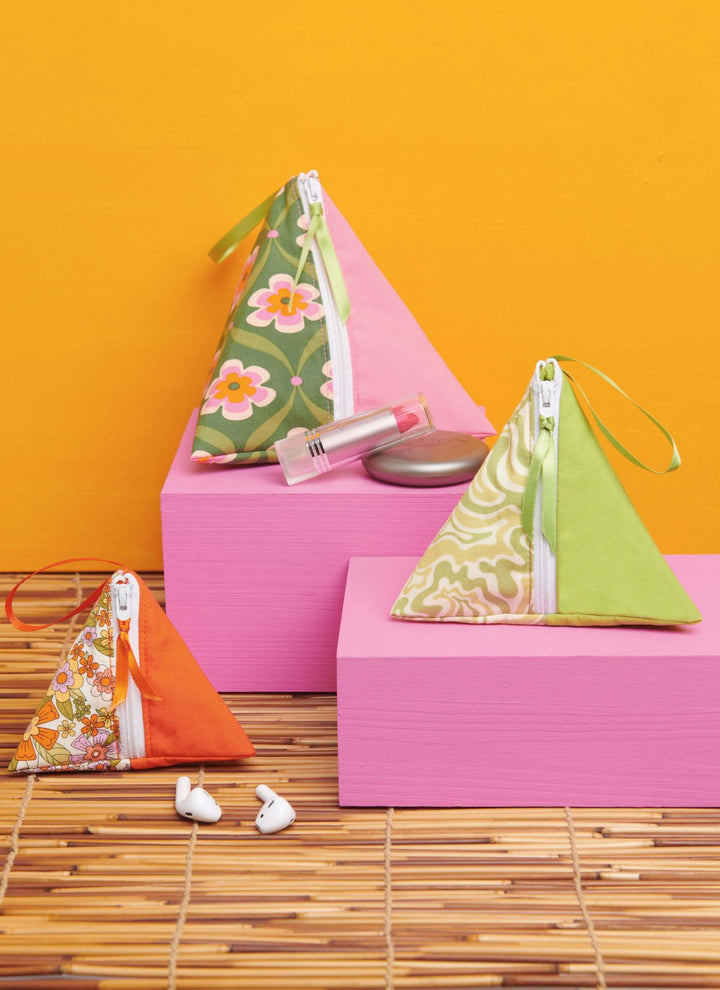 Displayed on pink blocks are three triangular fabric pouches in vibrant pink, green, and orange hues with white zippers. One pouch holds a lip balm and blush. Nearby, white earbuds sit on a bamboo mat against a bright orange background, perfectly complementing the playful essence of an empire waist top collection. These pouches are from the "Pattern - Simplicity - 9937- Hat, Tote Bag and Zipper Cases" by Super Cheap Fabrics.