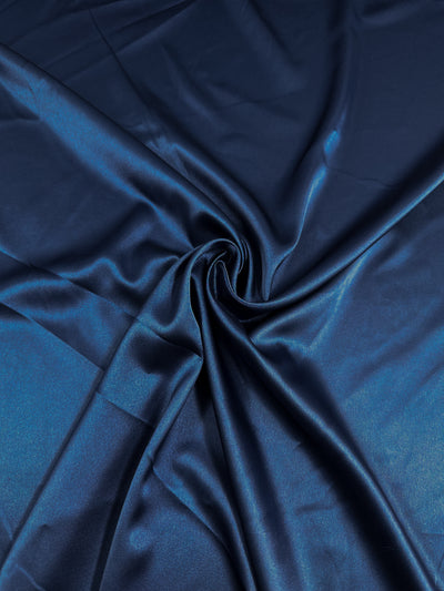 A close-up image of dark blue Satin Deluxe - Navy - 150cm fabric from Super Cheap Fabrics arranged in soft, smooth folds. The luxury fabric has a shiny, reflective surface that captures light and highlights its glossy texture. The material is gathered at the center, creating a swirl effect.
