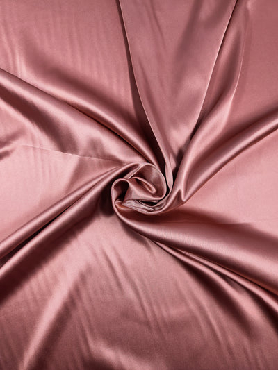 A close-up of a shiny, smooth pink Satin Deluxe - Canyon Clay - 150cm fabric from Super Cheap Fabrics with a soft shiny finish and light weight fabric. The soft folds radiate from a central twisted point, creating an elegant and luxurious texture.