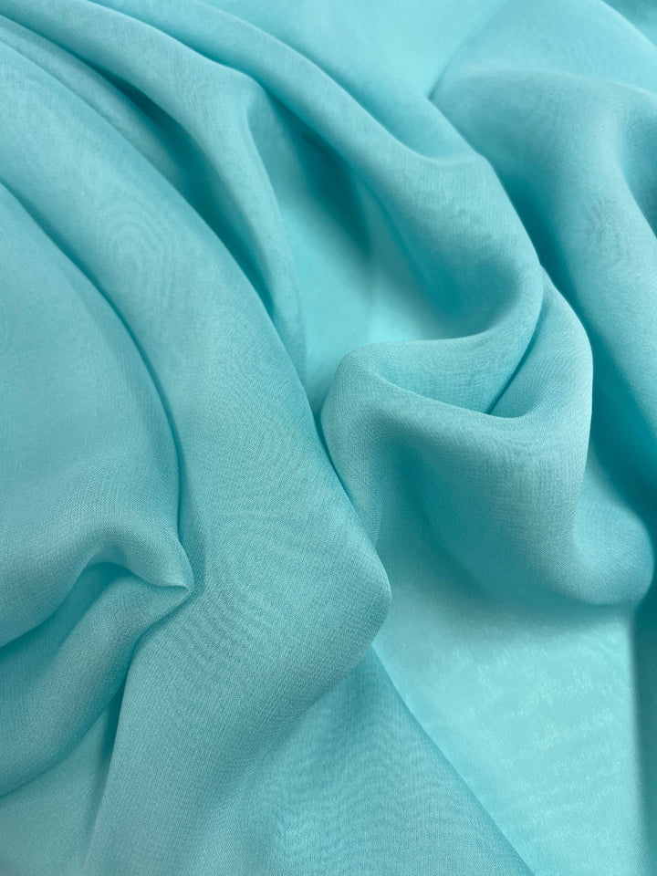 A close-up image of Super Cheap Fabrics' Silk Georgette - Tiffany - 135cm showcases its smooth and slightly crinkled texture. The folds create soft, flowing shapes and varying shades of light turquoise due to the light, making it perfect for spring and summer outfits.