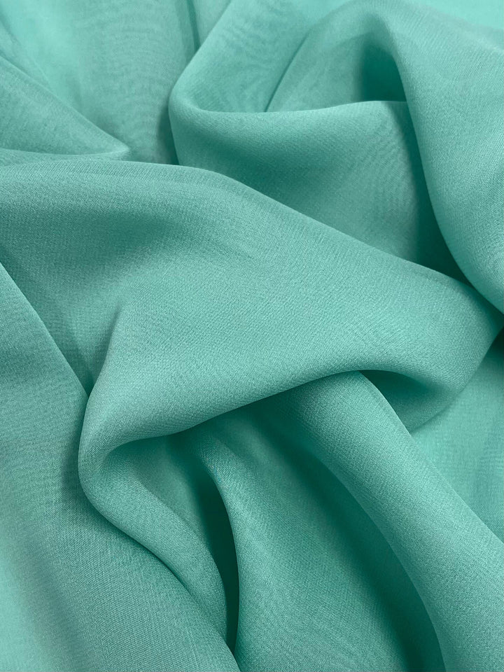 A close-up of the soft, light teal Silk Georgette - Lagoon from Super Cheap Fabrics, draped in gentle folds to create a smooth and textured appearance. This lightweight material offers a slight sheen, giving it an elegant look that's perfect for spring and summer outfits. The fabric measures 135cm in width.