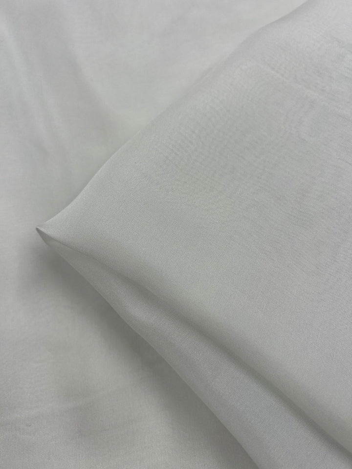 A close-up image of a pillow in platinum, made with Pure Silk Chiffon from Super Cheap Fabrics, resting on matching light gray bed sheets. The fabric appears smooth and soft, with subtle creases indicating the pillow's shape and the texture of the 100% silk material.