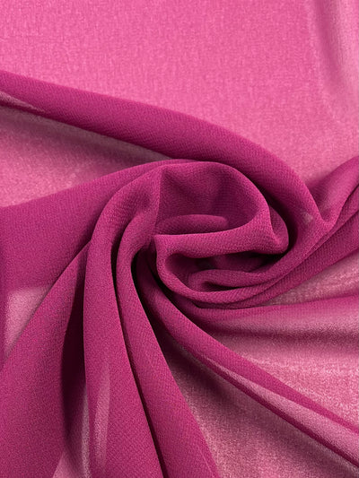 A close-up of a flowing, sheer, magenta-colored fabric intricately twisted and gathered at the center, showcasing its soft, lightweight texture and fine weave. Made from 100% polyester, the gentle folds and delicate transparency create an elegant, dynamic pattern perfect for floaty tops. This beautiful fabric is Hi-Multi Chiffon - Boysenberry - 150cm by Super Cheap Fabrics.