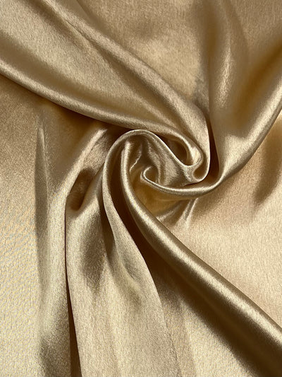 A close-up shot of Super Cheap Fabrics' luxurious Satin Back Crepe - Gold - 150cm. The smooth material, ideal for dressmaking, features a soft, silky texture and subtle sheen. Elegant folds and drapes create intricate shadows and highlights in this lightweight polyester marvel.
