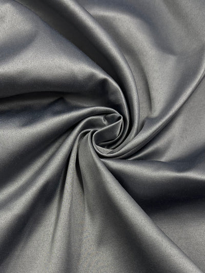 A close-up of smooth, Super Cheap Fabrics Delustered Satin - Ghost Gray - 150cm arranged in a spiral pattern. The material appears to be satin or silk, reflecting light and showcasing its glossy texture. The folds of the fabric create a dynamic and elegant visual effect, perfect for sophisticated workwear.