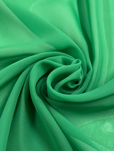 A close-up image of Super Cheap Fabrics' Hi-Multi Chiffon - Emerald - 150cm gathered in a circular, swirling pattern, creating a central focal point. The polyester fabric appears lightweight and slightly translucent, with subtle folds and texture visible throughout.