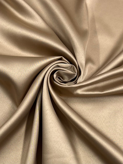 A close-up image of smooth, glossy beige Super Cheap Fabrics' Satin Deluxe - Gold - 150cm fabric arranged in a spiral pattern. The material has a slight sheen and appears silky and soft. The folds and swirls create a visually appealing texture, highlighting the light weight fabric's shiny finish.
