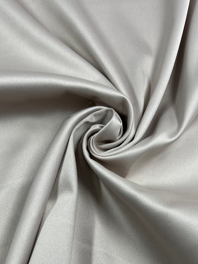 A close-up image of smooth, shiny, light gray medium weight fabric twisted into a swirling pattern. The texture appears soft and slightly reflective, showcasing gentle folds and curves—ideal for Delustered Satin - Tapioca - 150cm by Super Cheap Fabrics.