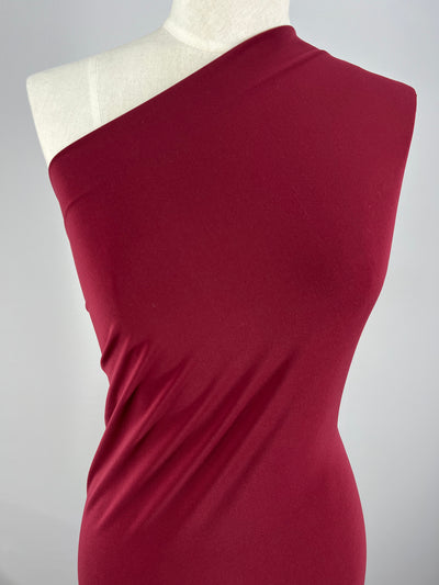 A close-up image of a mannequin dressed in a one-shoulder, form-fitting, maroon dress made from Super Cheap Fabrics' ITY Knit - Burgundy - 150cm. The dress has a sleek and smooth texture and drapes elegantly over the mannequin's body. The background is a plain light grey.
