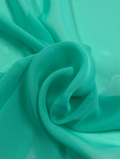 A close-up of a piece of Hi-Multi Chiffon - Florida Keys - 150cm in a bright turquoise color reminiscent of the Florida Keys. The 100% polyester material is gently gathered and draped in soft, flowing folds, creating a sense of movement and texture. The light shines through, highlighting its delicate nature. Available from Super Cheap Fabrics.