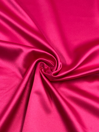 A close-up of Satin Deluxe - Hot Pink - 150cm from Super Cheap Fabrics, with a smooth and shiny finish. The fabric is slightly twisted in the center, creating soft folds and gentle curves. The light reflects off the surface, highlighting its glossy and silky appearance.