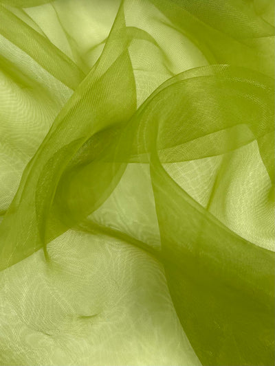 A close-up of translucent, flowing green fabric, creating a dynamic and fluid composition. The extra lightweight Organza - Going Green - 150cm by Super Cheap Fabrics appears light and airy, with soft folds and gentle curves that catch and diffuse the light, giving a sense of movement and texture.