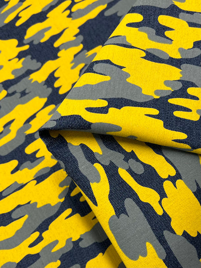 A piece of medium to heavy weight fabric with a camo pattern featuring yellow, grey, and black colors, folded in the center. The design has an abstract, wavy appearance resembling traditional camouflage. Made from 100% cotton, it is best maintained with a cold wash. This is the Printed Denim - Camouflage - 145cm by Super Cheap Fabrics.
