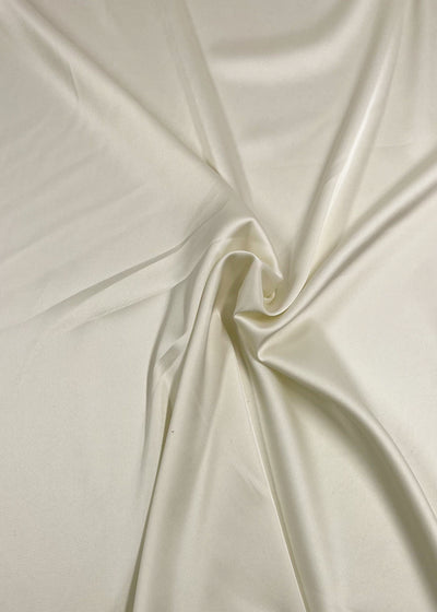 A close-up image of a piece of Satin Deluxe - Ivory - 150cm by Super Cheap Fabrics that appears to have a soft, shiny finish. The material is slightly wrinkled and gathered in the center, creating gentle folds and smooth textures throughout. The satin reflects light beautifully, giving it a glossy appearance.