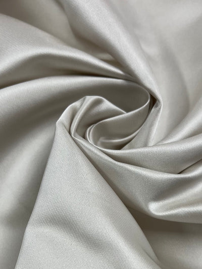 Close-up of a piece of smooth, shiny, silver-grey Delustered Satin - Gardenia - 150cm. The Super Cheap Fabrics polyester fabric is arranged with gentle folds and swirls, creating a soft and elegant appearance. The texture appears silky and delicate, reflecting light subtly.