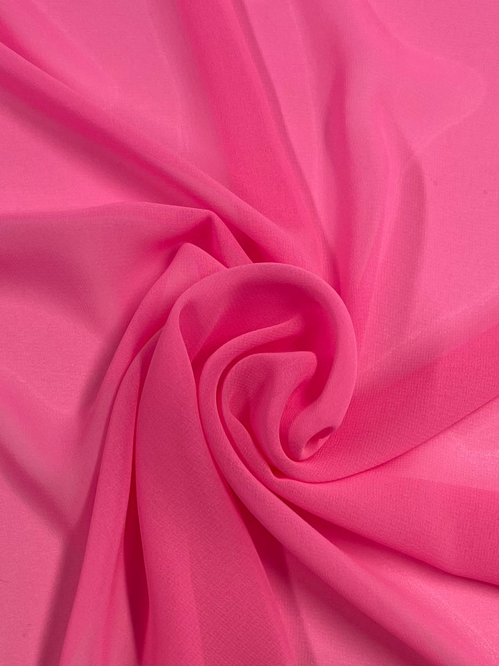 A piece of Super Cheap Fabrics' Hi-Multi Chiffon - Fluoro Pink - 150cm fabric is shown in the image. The material is softly draped and twisted into a gentle spiral pattern, creating smooth and flowing folds. The texture appears lightweight, sheer, and slightly translucent, emphasizing its 100% polyester composition.