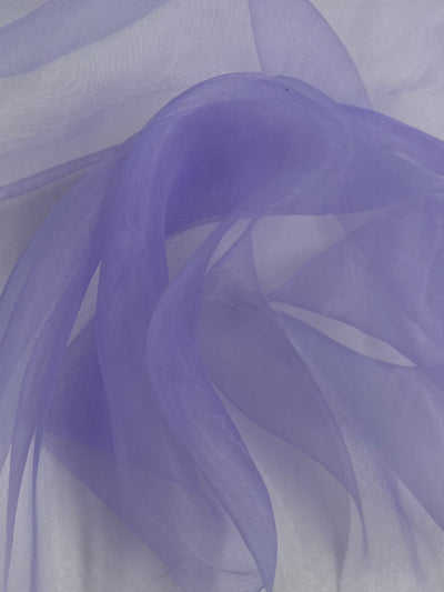 A close-up image of smooth, sheer Organza - Lavender - 150cm fabric arranged in soft, flowing curves. The translucent nylon material from Super Cheap Fabrics creates a delicate, ethereal effect with varying shades of purple as the light filters through. The background is an out-of-focus gradient of similar tones.