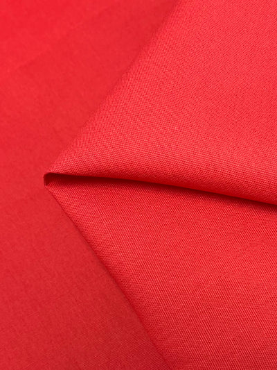 A close-up image of Super Cheap Fabrics' Linen Canvas - Hibiscus - 150cm with a smooth, fine texture. The medium-weight fabric is neatly folded, creating a sharp, triangular crease in the center of the image. The vibrant hibiscus red color is consistent throughout the fabric.