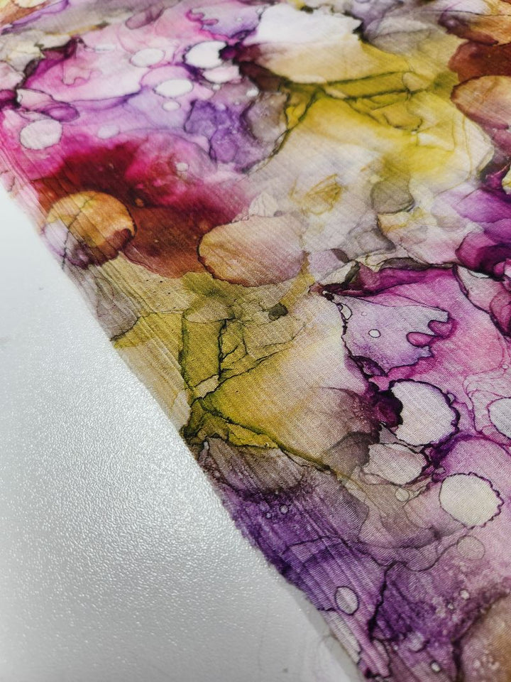 A close-up of the Designer Cotton - Blist from Super Cheap Fabrics showcases an abstract watercolor pattern with vibrant pink, yellow, purple, and green swirls and spots. The breathable fabric's texture and dynamic flow of colors form an artistic and fluid design, ideal for luxury sewing projects.