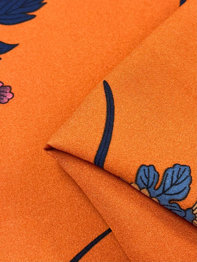 Close-up of Super Cheap Fabrics' Printed Crepe - Tuscan Flor (150cm), displaying a folded high-fashion orange fabric with blue and purple floral patterns. This lightweight polyester offers a smooth, slightly glossy texture.