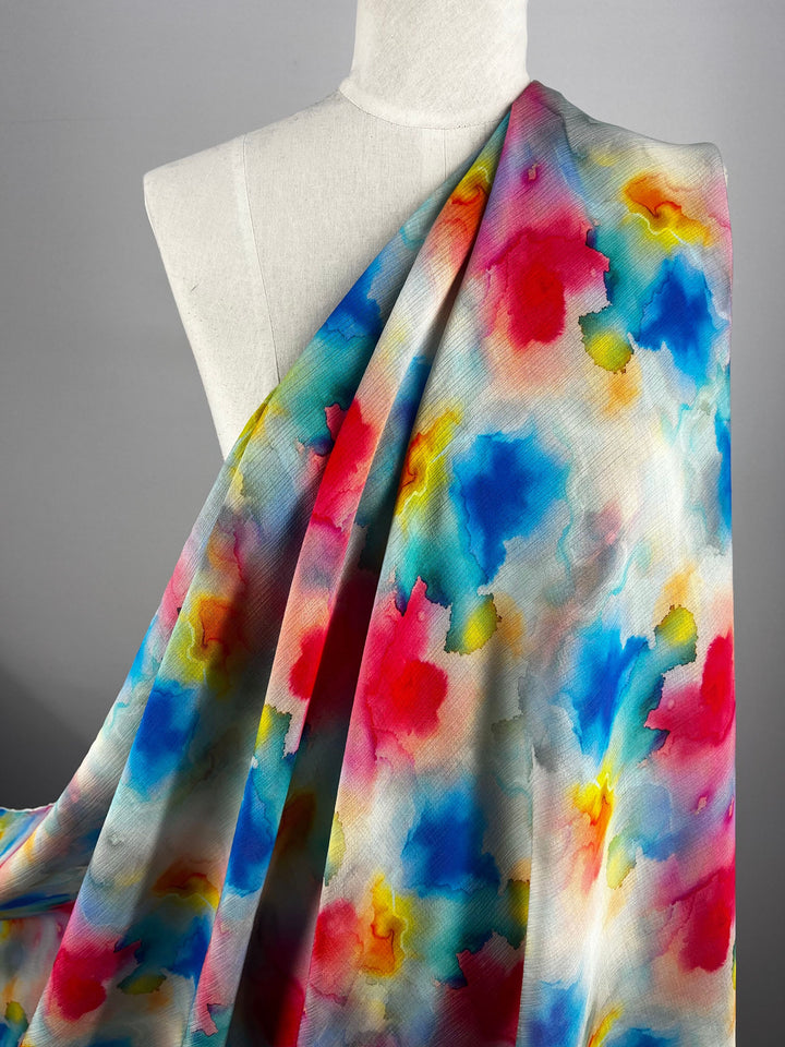 A mannequin is draped with a stunning Designer Cotton from the Venus collection by Super Cheap Fabrics. This luxurious fabric boasts a colorful abstract pattern of pink, blue, yellow, and orange splashes, reminiscent of watercolor art—ideal for upscale sewing projects. The fabric measures 150 cm wide.