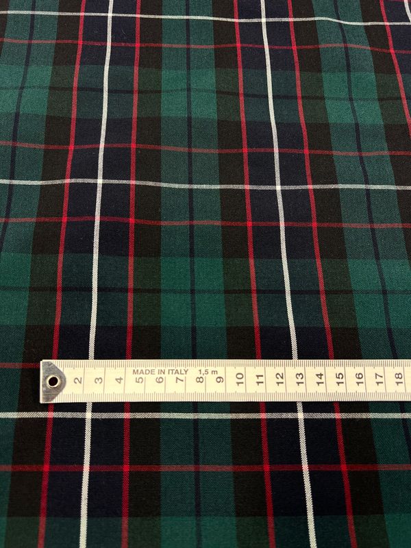 A detailed view of the Shirting - Hills - 145cm by Super Cheap Fabrics showcases a lightweight polyester rayon fabric in green, navy, and red plaid. A white measuring tape laid horizontally across it indicates measurements up to 15 centimeters, while the intersecting lines create perfect squares in this versatile multiuse textile.