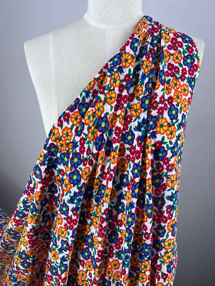 A Super Cheap Fabrics mannequin elegantly displays the Salt Shrunk Print - Pansy, a single-shoulder draped fabric with a stunning floral pattern in red, orange, yellow, blue, and green on a classic white background.