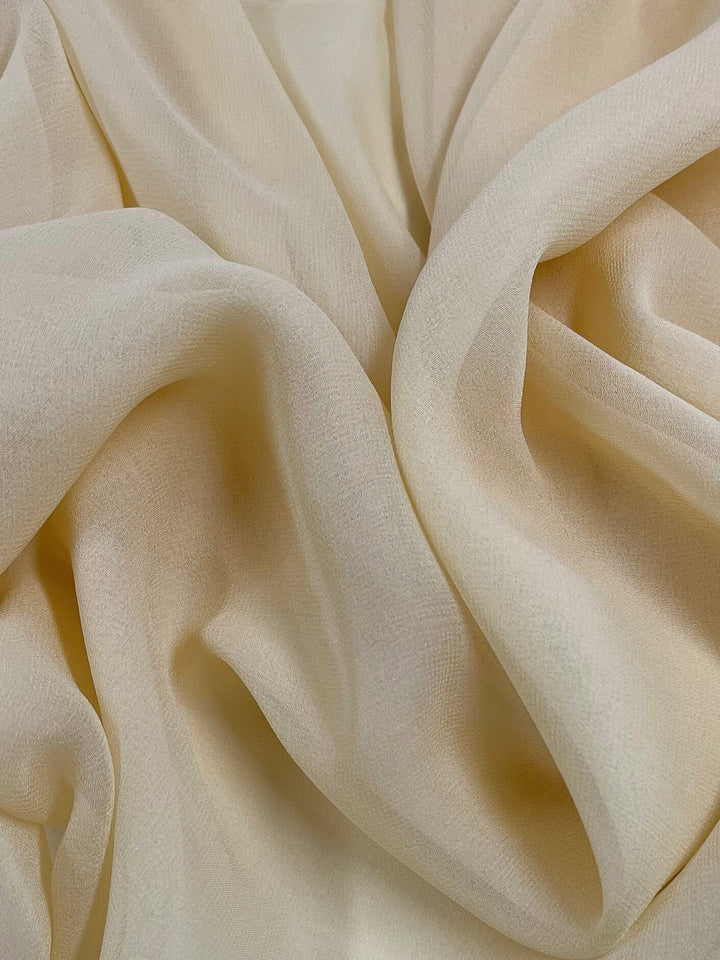 A close-up view of Super Cheap Fabrics' Silk Georgette - Chardonnay - 135cm reveals a soft, cream-colored plain silk fabric with gentle folds and drapes. The lightweight material appears smooth and delicate, forming fluid and wavy patterns. The lighting enhances the texture and subtle shading, making it perfect for elegant summer outfits.