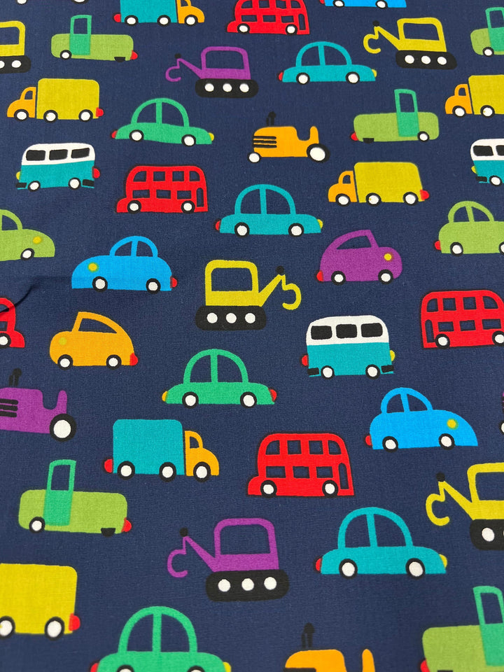 Super Cheap Fabrics' Printed Cotton - Mobile - 110cm features a vibrant pattern of cartoon vehicles like cars, trucks, and buses on navy blue. Made from natural cotton fibers in bright yellow, red, green, and blue hues, this versatile fabric is ideal for a range of uses.