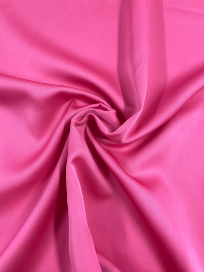 A piece of Habutae Satin - Azalea Pink - 150cm from Super Cheap Fabrics is elegantly twisted and draped. The lightweight material appears smooth and soft, with gentle folds creating a swirl pattern in the center. The lighting highlights the rich color and texture of this polyester fabric.