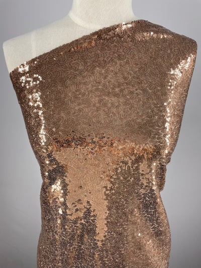 A close-up of a torso mannequin draped with champagne-colored sequin fabric. The Super Cheap Fabrics' Evening Sequins - Cafe - 150cm, ideal for festival wear, has a shiny, reflective surface with a shimmering appearance. The draping creates a one-shoulder look, emphasizing the Evening Sequins - Cafe's texture and sparkle.