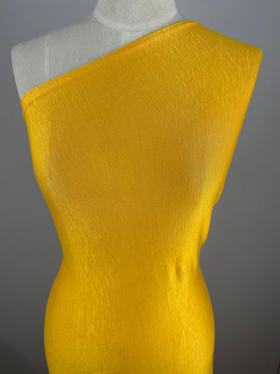 A mannequin is adorned with a vivid yellow one-shoulder dress made from Super Cheap Fabrics' Bamboo Jersey - Gold Fusion - 165cm. The material appears textured, adding subtle detail to the smooth and form-fitting design. The background is plain and grey, ensuring the focus remains entirely on this environmentally responsible dress.