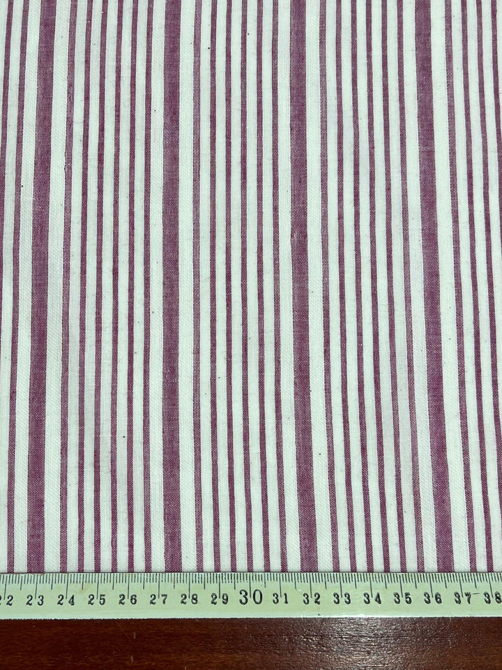 A piece of Super Cheap Fabrics' Cotton Voile - Magenta - 150cm with alternating thin and thick purple vertical stripes on a white background. A green measuring tape placed at the bottom shows measurements in centimeters, indicating the scale of the fabric. The lightweight fabric is laid on a brown wooden surface.